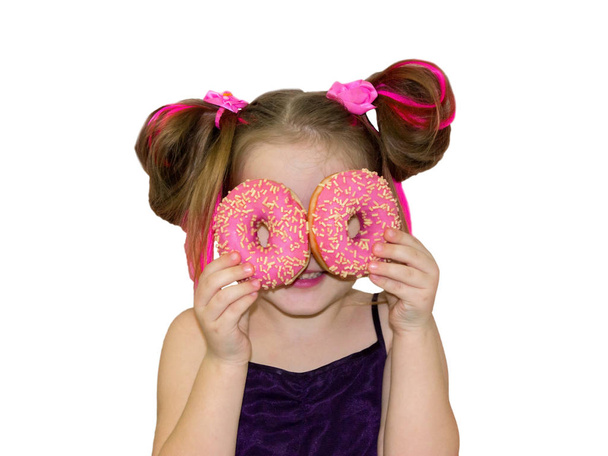A little kid girl dabbles and plays with two fresh donuts before eating. A child holds donuts near his eyes and looks through the holes like through glasses. - Foto, imagen