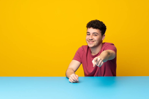 Молодой человек с красочной стеной и столом указывает пальцем на вас с уверенным выражением лица
 - Фото, изображение