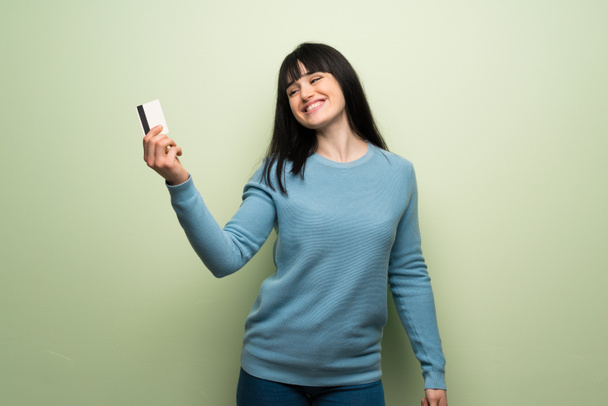 Young woman over green wall holding a credit card and thinking - Photo, Image