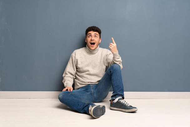 Mladý muž sedí na podlaze směřující nahoru a překvapený - Fotografie, Obrázek