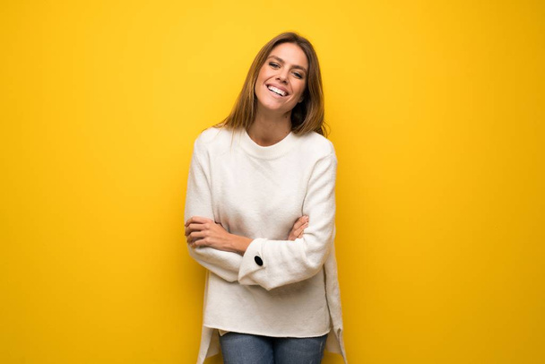 Mujer rubia sobre pared amarilla manteniendo los brazos cruzados mientras sonríe
 - Foto, Imagen