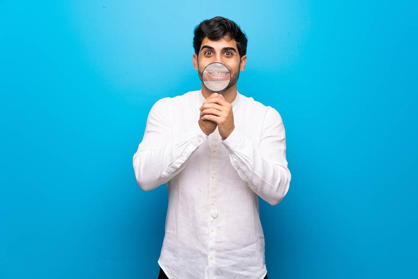 Νέος άνθρωπος πάνω από απομονωμένο μπλε τοίχο παίρνοντας ένα μεγεθυντικό φακό και δείχνοντας τα δόντια του - Φωτογραφία, εικόνα