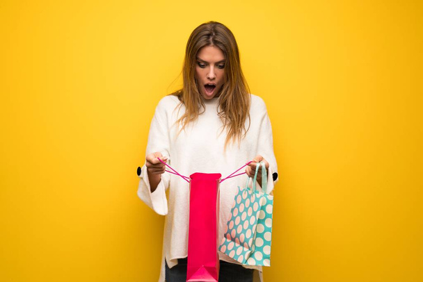 Blonde woman over yellow wall surprised while holding a lot of shopping bags - 写真・画像