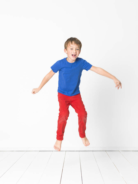 cute boy in blue t-shirt and red trousers jumping high over wooden floor in front of white wall in studio  - Fotó, kép