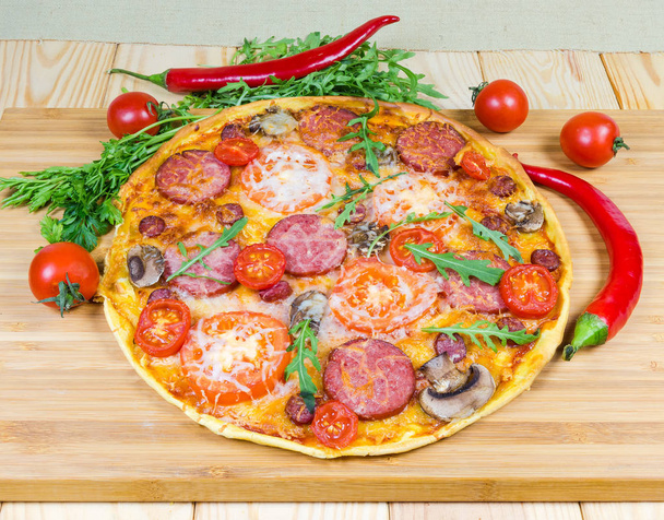 Pizza with sausages and mushrooms on cutting board among vegetables - Photo, Image