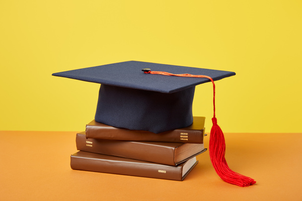 Academic cap and brown books on orange surface isolated on yellow - Foto, imagen