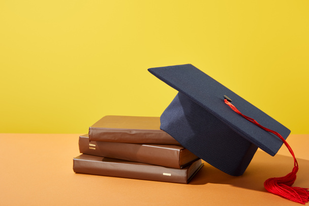 Brown books and academic cap with red tassel on orange surface isolated on yellow - Photo, Image