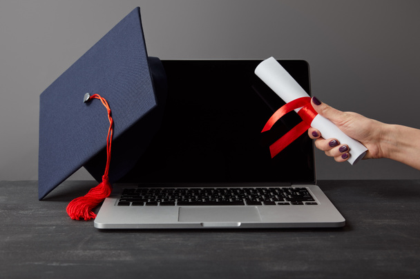Cropped view of woman holding diploma near laptop with blank screen and academic cap on grey - Photo, Image