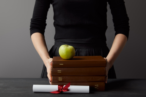 Cropped view of woman holding brown books and green apple near diploma on grey - Photo, Image