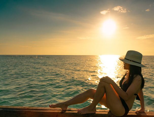 Sexy and relax woman wear black bikini with straw hat sit on wooden beam near sand beach at sunset. Girl enjoy holiday at tropical paradise beach on summer vacation. Holiday travel. Summer vibes. - Photo, Image