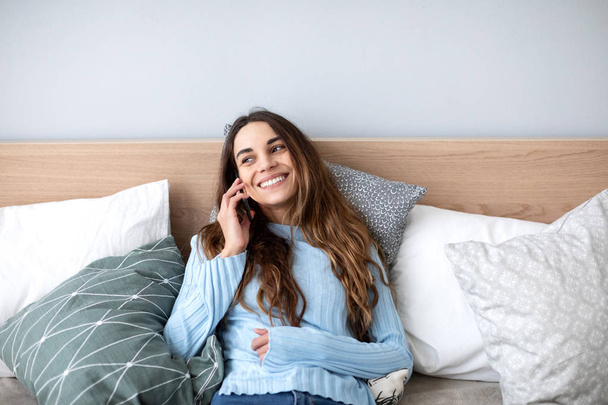 Beautiful smiling woman talking on cellphone while lying in cozy bed at home. - 写真・画像