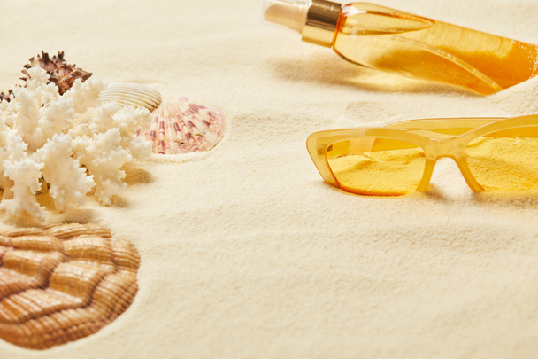 selective focus of seashell near white coral, yellow sunglasses and suntan oil bottle on sand  - Fotó, kép
