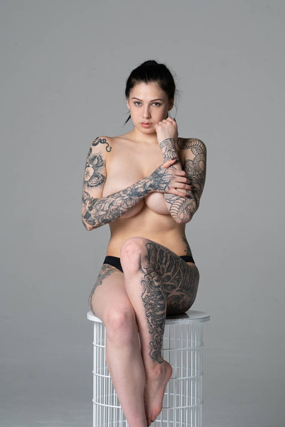 young beautiful girl with tattoo posing nude in studio sitting on a chair in black underwear - Фото, изображение