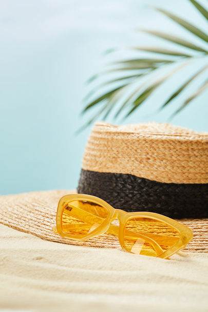 selective focus of yellow sunglasses near straw hat on golden sand in summertime isolated on blue - Photo, Image