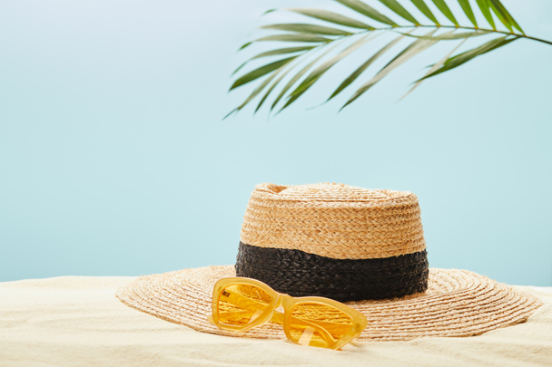 selective focus of yellow sunglasses near straw hat on sand in summertime isolated on blue - Photo, Image