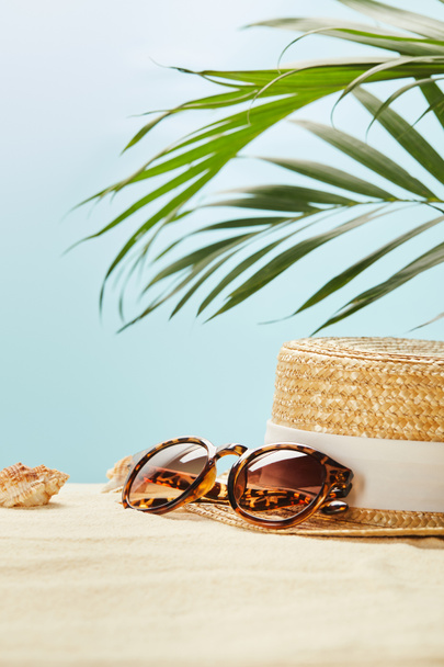 selective focus of sunglasses near straw hat and seashells in summertime isolated on blue - Фото, изображение