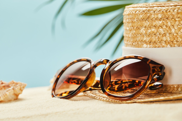 selective focus of sunglasses and straw hat near seashell in summertime isolated on blue - Photo, Image