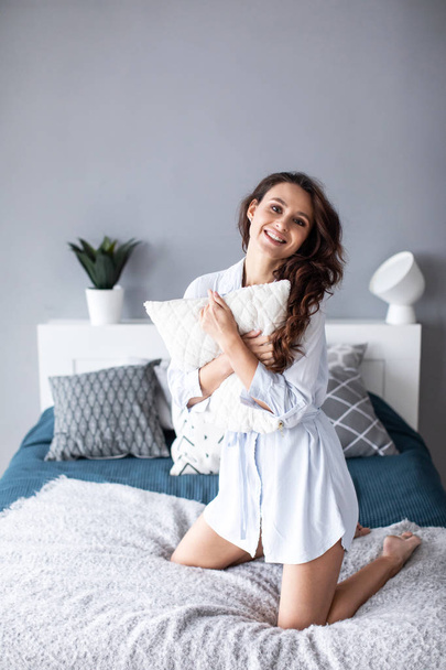 Positive woman with pillow sitting in bed and smiling to camera at home.  - Foto, Imagen