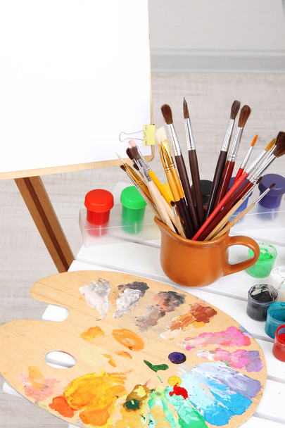 Wooden easel with clean paper and art supplies in room - Фото, изображение