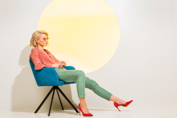 beautiful fashionable girl in colorful clothes sitting in armchair on white with yellow circle and copy space - Photo, Image