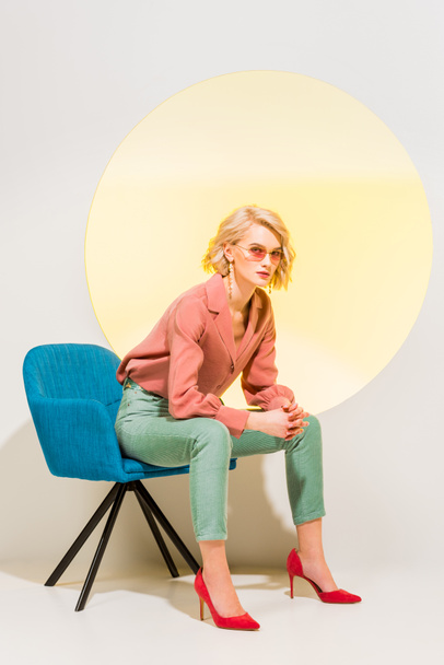 beautiful fashionable girl in colorful clothes sitting in armchair on white with yellow circle - Φωτογραφία, εικόνα