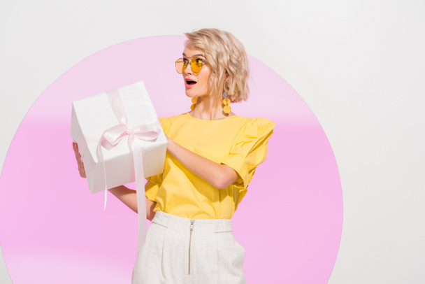 excited stylish girl holding gift box on white with pink circle - Foto, Imagem
