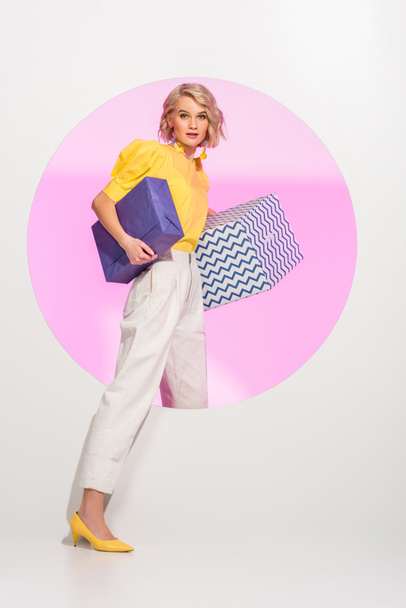beautiful fashionable girl holding gift boxes, looking at camera and posing on white with pink circle - Foto, Imagem