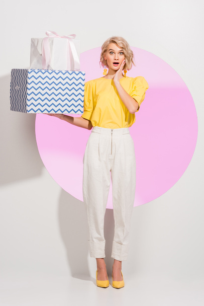 surprised fashionable girl gesturing and holding gift boxes on white with pink circle - Foto, imagen