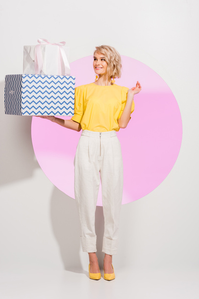 beautiful fashionable girl holding gift boxes and smiling on white with pink circle - Φωτογραφία, εικόνα