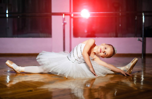 Beautiful preschool girl in a classical ballet tutu stretching her body on the wooden studio floor. Pretty little ballerina doing exercises on the floor and looking at camera. - Fotoğraf, Görsel