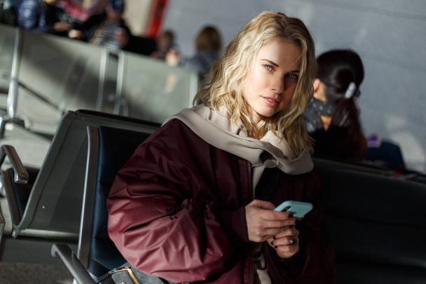 Curly blonde with phone in hands sitting at waiting room. - Photo, Image