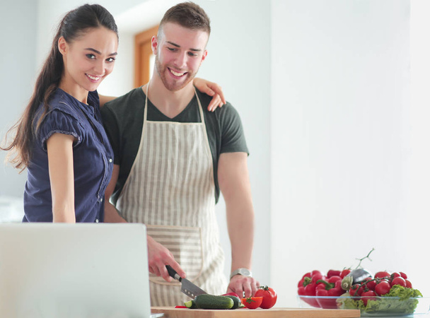Young man cutting vegetables and woman standing with laptop in the kitchen - Фото, изображение