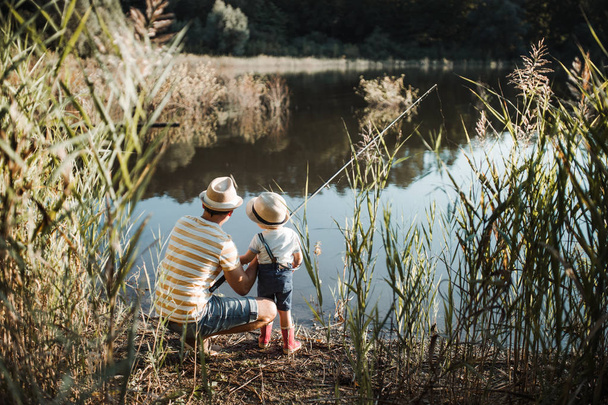 A rear view of mature father with a small toddler son outdoors fishing by a lake. - Foto, Imagem
