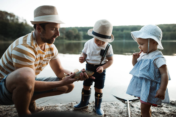 A mature father with small toddler children fishing by a lake, holding a fish. - Foto, Imagem
