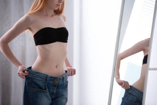 Skinny girl trying on too big jeans in front of a mirror - Foto, imagen