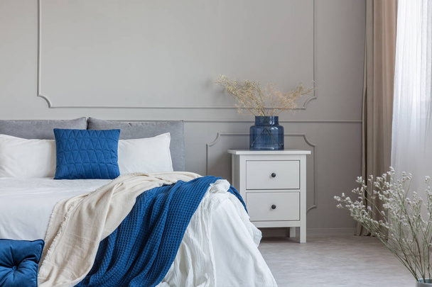 Flowers in blue glass vase on white wooden nightstand with drawers next to king size bed with blue white and grey bedding - Foto, afbeelding