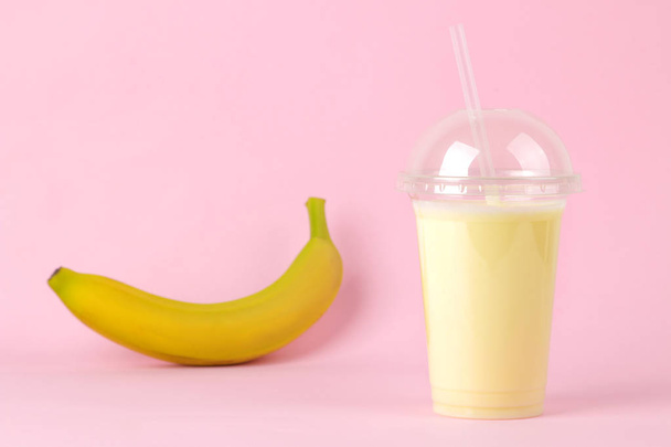Haciendo un batido. un vaso desechable de plástico con un batido de plátano y un plátano fresco sobre un fondo rosa brillante de moda
. - Foto, Imagen