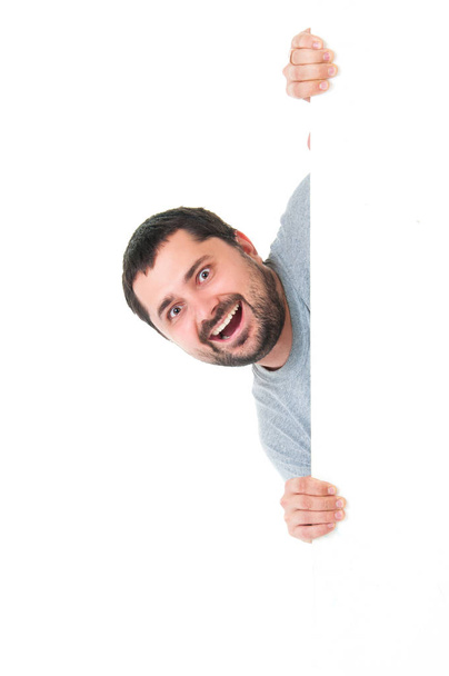 Homem barbudo bonito sorrindo vestindo uma camiseta azul cinza e posando atrás de um painel branco isolado no fundo branco. Lugar para publicidade
 - Foto, Imagem