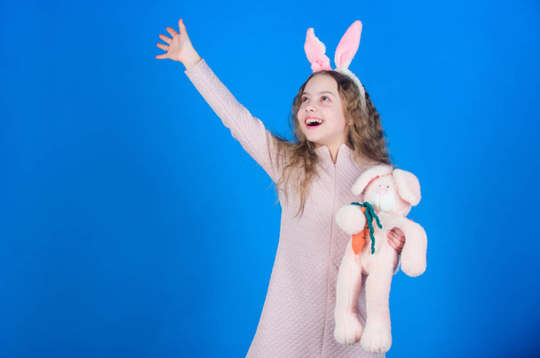 Child smiling play bunny toy. Happy childhood. Get in easter spirit. Bunny ears accessory. Lovely playful bunny child with long hair. Cute and adorable. Bunny girl with cute toy on blue background - Fotó, kép