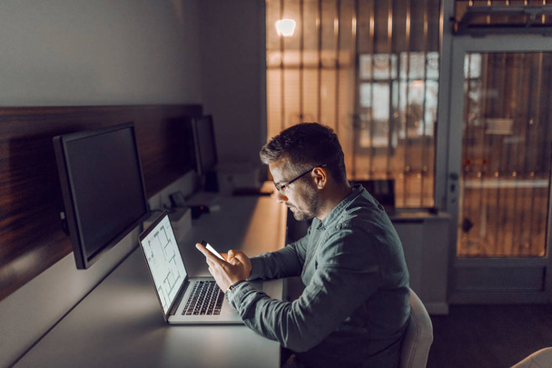 Sivunäkymä nuori valkoihoinen ahkera arkkitehti silmälasit älypuhelimella istuessaan toimistossa myöhään yöllä. Hänen edessään kannettava tietokone
. - Valokuva, kuva
