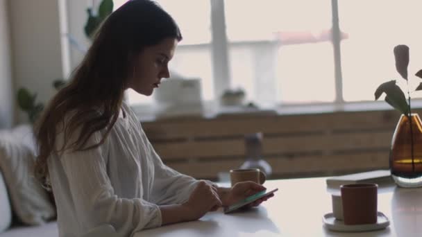 Κομψό κορίτσι κρατώντας smartphone καθιστό εσωτερικό καφέ με οπίσθιο φωτισμό 4K πλευρική θέα. Πορτρέτο της νεαρής γυναίκας που πληκτρολογεί κινητό τηλέφωνο στο διάλειμμα καφέ φυσικό φως άνετο εσωτερικό. Σύγχρονες τεχνολογίες κοινωνικά δίκτυα - Πλάνα, βίντεο