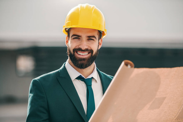 Portrait of handsome smiling successful architect in formal wear and with helmet holding plans while standing on site. You have to work for success, not dream about it. - Photo, Image