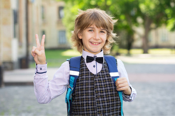 Boy schoolboy with notebook and books on the background of the college shows a sign V. Complete the school year and go on vacation. Or back to school in second grade. - Zdjęcie, obraz