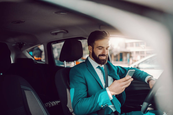 Young Caucasian successful bearded businessman in turquoise suit and with toothy smile sitting in the car and using smart phone. The expert in anything was once a beginner. - Valokuva, kuva