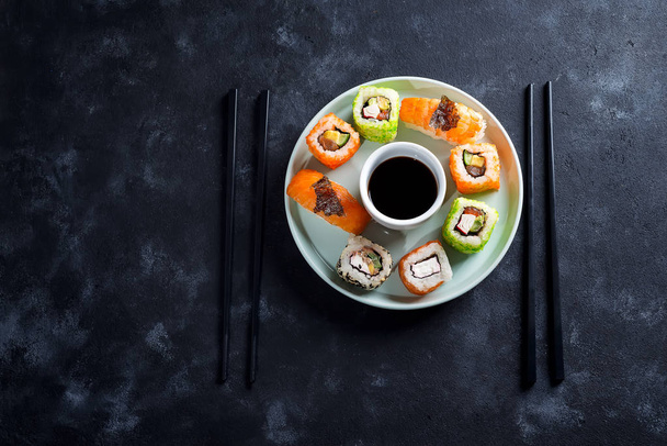 Різні свіжі та смачні суші, встановлені на керамічній тарілці зі сланцевими паличками, соус на фоні чорного каменю, плоска покладена
 - Фото, зображення