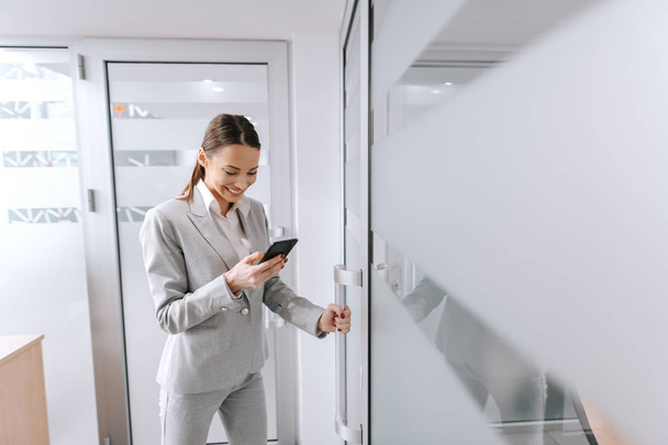 Jovem CEO do sexo feminino caucasiano vestido de cinza formal desgaste entrar no escritório e usando telefone inteligente. Para ser um bom líder leva tempo e experiência
. - Foto, Imagem