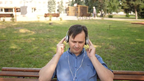 joven guapo escuchando música desde su teléfono inteligente con auriculares, bailando afuera en el parque en un banco en el centro de Europa
 - Foto, Imagen