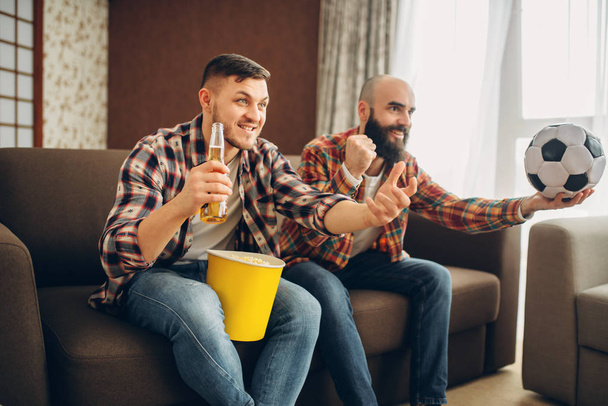 Two male football fans wathing tv broadcast at home. Friends with beer and popcorn cheer for their favorite team, decisive match - Photo, image
