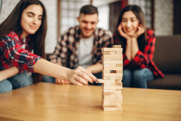 Smiling friends plays jenga at home, selective focus on tower. Board game with wooden blocks requiring high concentration, entertainment for funny company - Valokuva, kuva