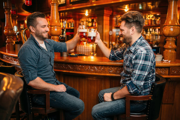 Dois amigos sorridentes bebem cerveja no balcão do pub. Homens barbudos com canecas se divertindo no bar, bons amigos
 - Foto, Imagem